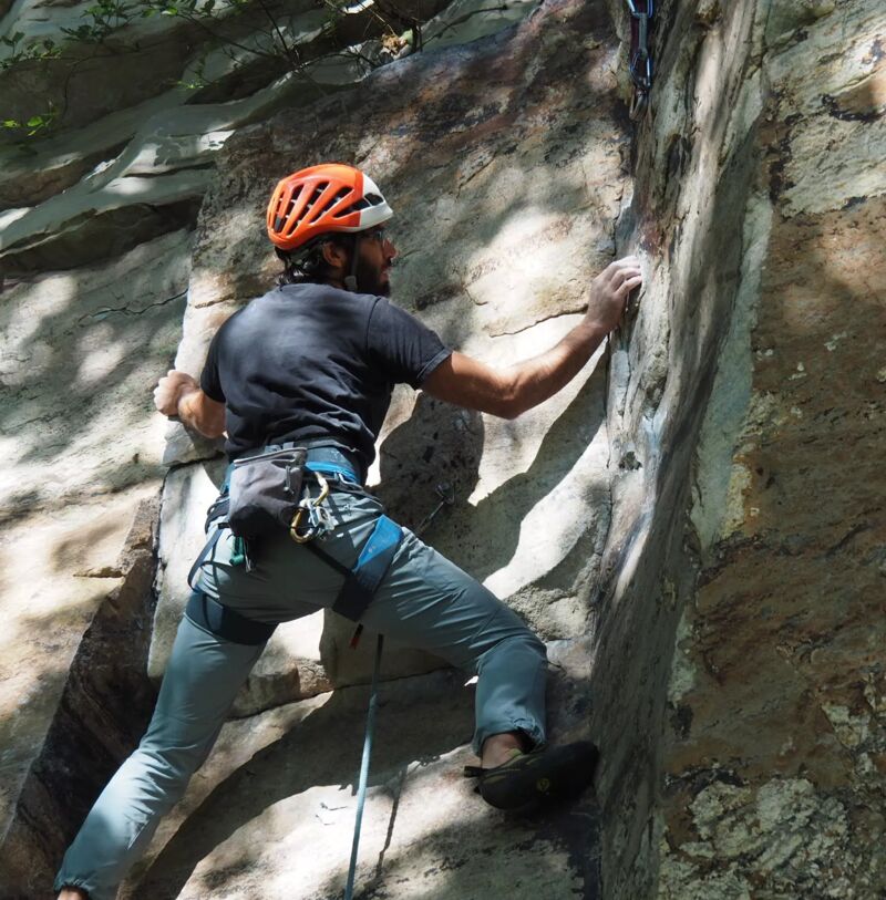 A man is rock climbing on a sunny day. He is wearing a helmet, a harness, and climbing shoes. He is reaching for a hold with his right hand. The rock face is steep and textured. The man appears focused and determined as he ascends the rock. Shadows from the surrounding trees create contrast on the rock face, highlighting the climber's form and the challenging nature of the climb.